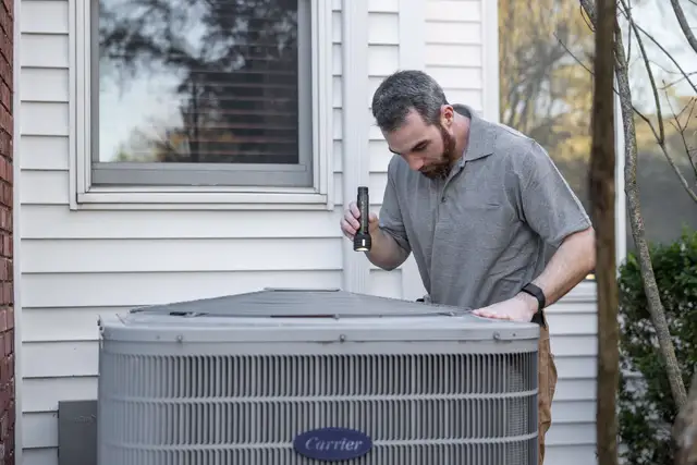 Technician checking the outdoor unit of a heat pump beside a white clapboard house